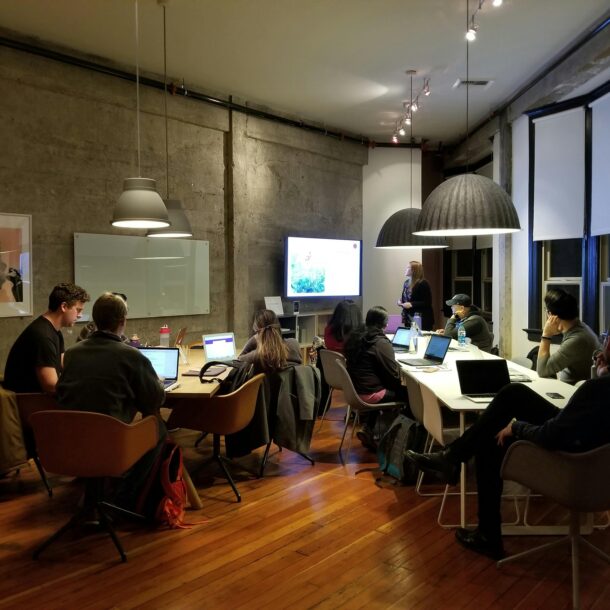 Group of people in a modern conference room attending a presentation. Some are seated around a large table with laptops open, while a woman stands at the front near a screen displaying a slide. The room features industrial-style walls, large hanging lamps, and wooden floors.