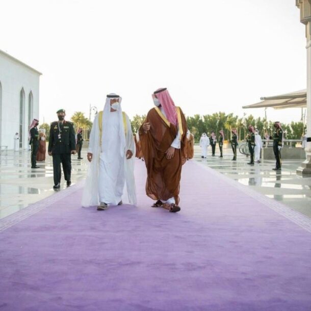 Two Gulf leaders walk side by side on a long purple carpet during a formal welcoming ceremony, flanked by uniformed guards in dark green with red sashes and keffiyehs. The architecture is grand and symmetrical, with white marble columns and polished stone. The use of purple underfoot reflects prestige and ceremonial power, while the red accents worn by guards symbolize strength and honor, highlighting the deep cultural symbolism of red and purple in diplomatic protocol.