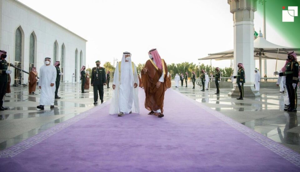 Two Gulf leaders walk side by side on a long purple carpet during a formal welcoming ceremony, flanked by uniformed guards in dark green with red sashes and keffiyehs. The architecture is grand and symmetrical, with white marble columns and polished stone. The use of purple underfoot reflects prestige and ceremonial power, while the red accents worn by guards symbolize strength and honor, highlighting the deep cultural symbolism of red and purple in diplomatic protocol.