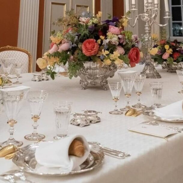 Elegant formal dining table with folded napkins resting on silver plates, surrounded by crystal glassware, floral centerpieces, and gold-accented cutlery.