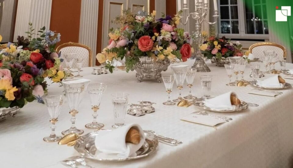 Elegant formal dining table with folded napkins resting on silver plates, surrounded by crystal glassware, floral centerpieces, and gold-accented cutlery.