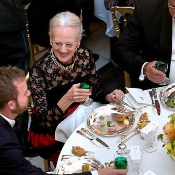 Formal dining event with well-dressed guests seated at a round table, enjoying a meal in perfect rhythm. Silver cutlery, white linens, and floral centerpieces enhance the elegant ambiance.