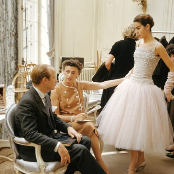 1950s haute couture fitting session inside a Parisian fashion house. A model in a pearl-studded gown poses elegantly, as seated designers and clients observe and discuss details. The setting is refined, with floral curtains, soft lighting, and fashion sketches around the room.