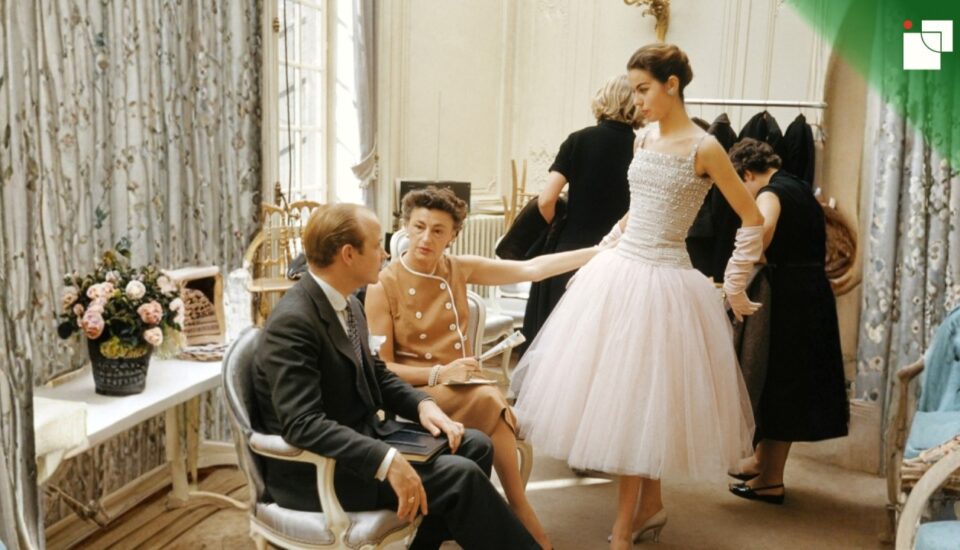 1950s haute couture fitting session inside a Parisian fashion house. A model in a pearl-studded gown poses elegantly, as seated designers and clients observe and discuss details. The setting is refined, with floral curtains, soft lighting, and fashion sketches around the room.