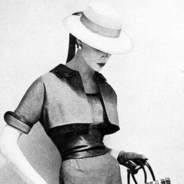 Vintage black-and-white fashion photo of a woman wearing a tailored dress, leather belt, wide-brimmed hat, and gloves. She holds a small handbag and looks downward, capturing timeless elegance and understated sophistication.