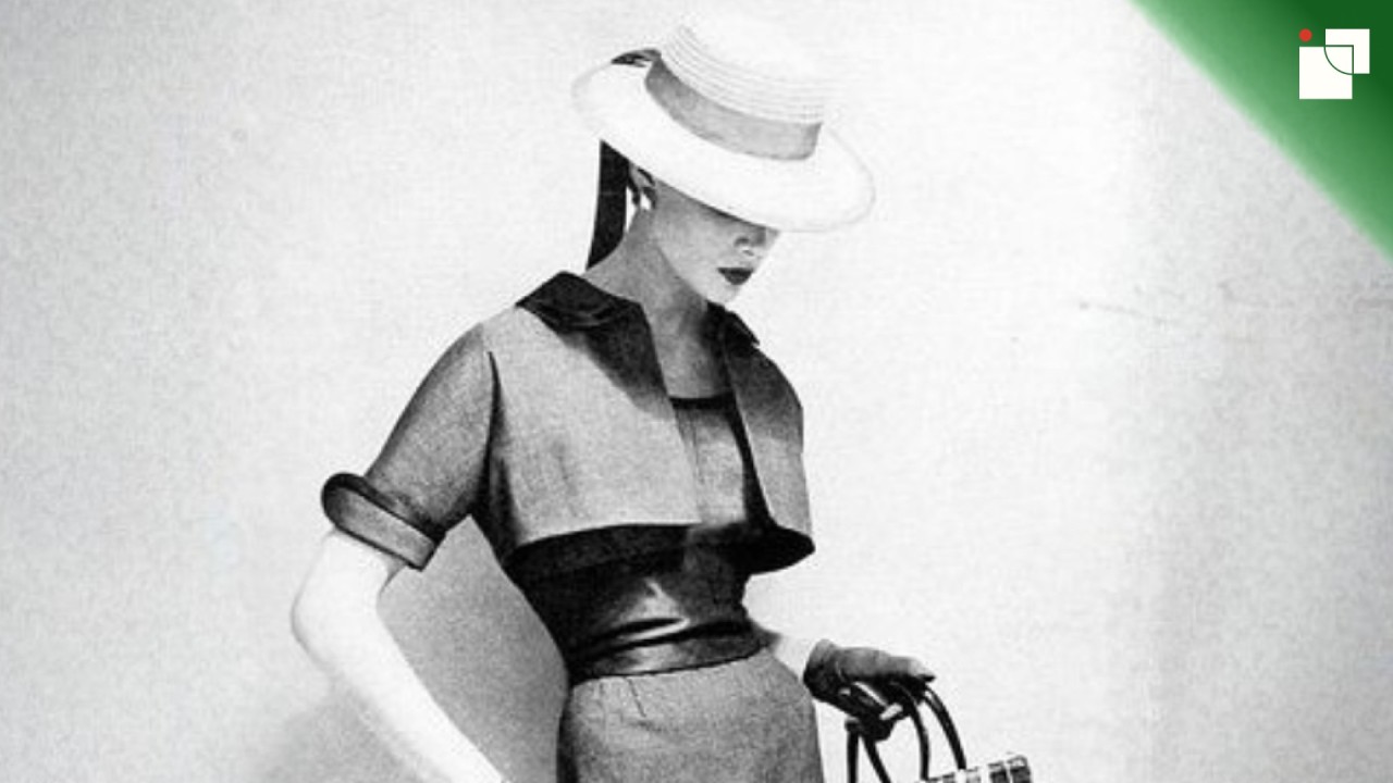 Vintage black-and-white fashion photo of a woman wearing a tailored dress, leather belt, wide-brimmed hat, and gloves. She holds a small handbag and looks downward, capturing timeless elegance and understated sophistication.