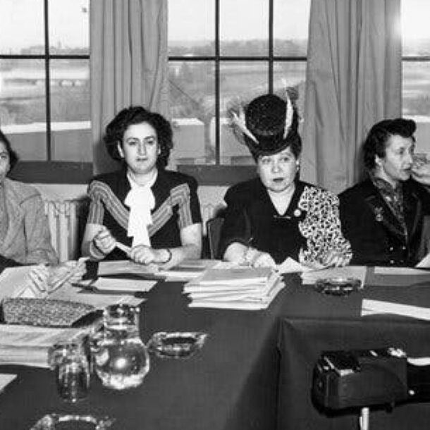 Black-and-white photo of seven influential women seated around a table during a mid-20th-century diplomatic meeting. They wear formal attire, some with hats and traditional dress, representing early female involvement in international diplomacy.